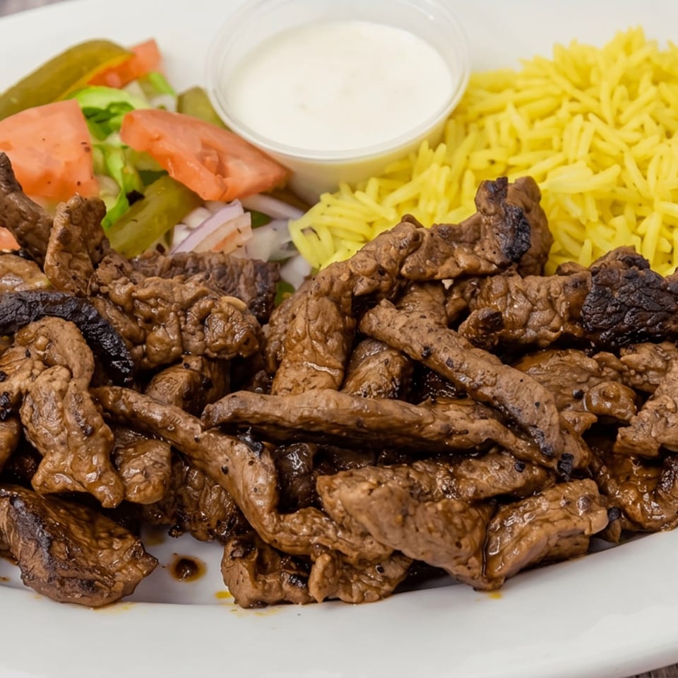 Grilled beef strips served with yellow rice, salad, pickles, tomato, and a side of creamy sauce on a white plate.